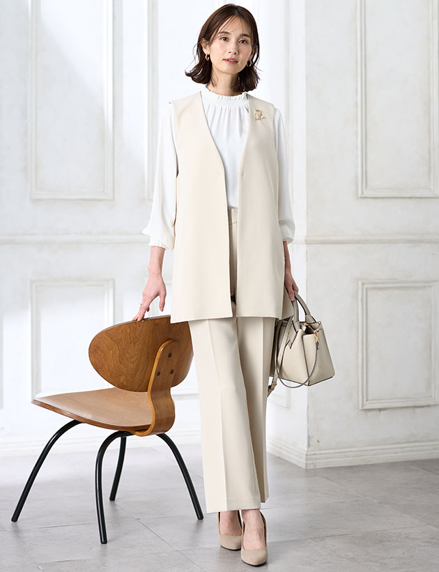 アイボリーのジレジャケット【セットアップ対応】・アイボリーのストレートパンツ【セットアップ対応】・ホワイトのフリルブラウス・アイボリーのバイカラーバッグを着用した女性の画像