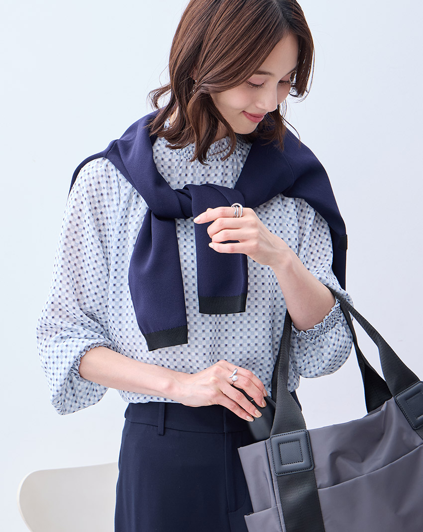 サックスブルーのジャガードTブラウス・ネイビーの【アルファブリック】ワイドパンツ【セットアップ対応】・ネイビーのバイカラーニットカーディガン【ハンドウォッシャブル】・チャコールグレーの軽量A4トートバッグを着用した女性のアップの画像