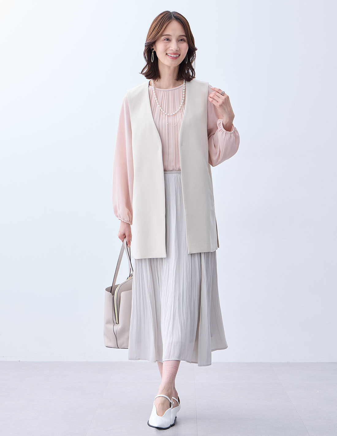 ベージュのジレジャケット【セットアップ対応】・ピンクのプリーツTブラウス・ライトグレーのプリーツスカート・パールネックレスを着用した女性の画像