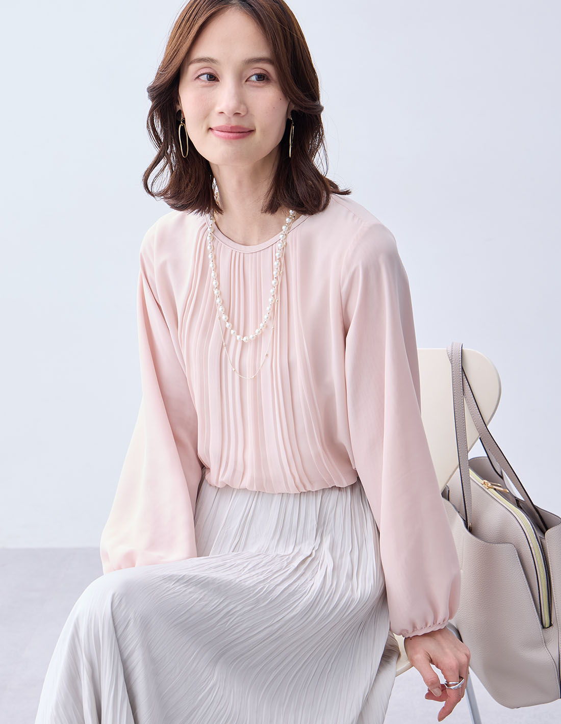 ピンクのプリーツTブラウス・ライトグレーのプリーツスカート・パールネックレスを着用した女性のアップの画像