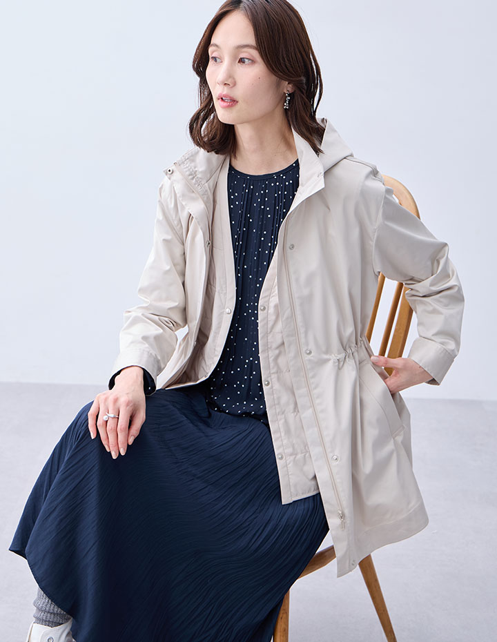 ライトベージュの3WAYコート【スーパーウォーム】【ライナー付き】・ブラックのジャガードTブラウス・ネイビーのプリーツスカートを着用した女性が椅子に座っている画像