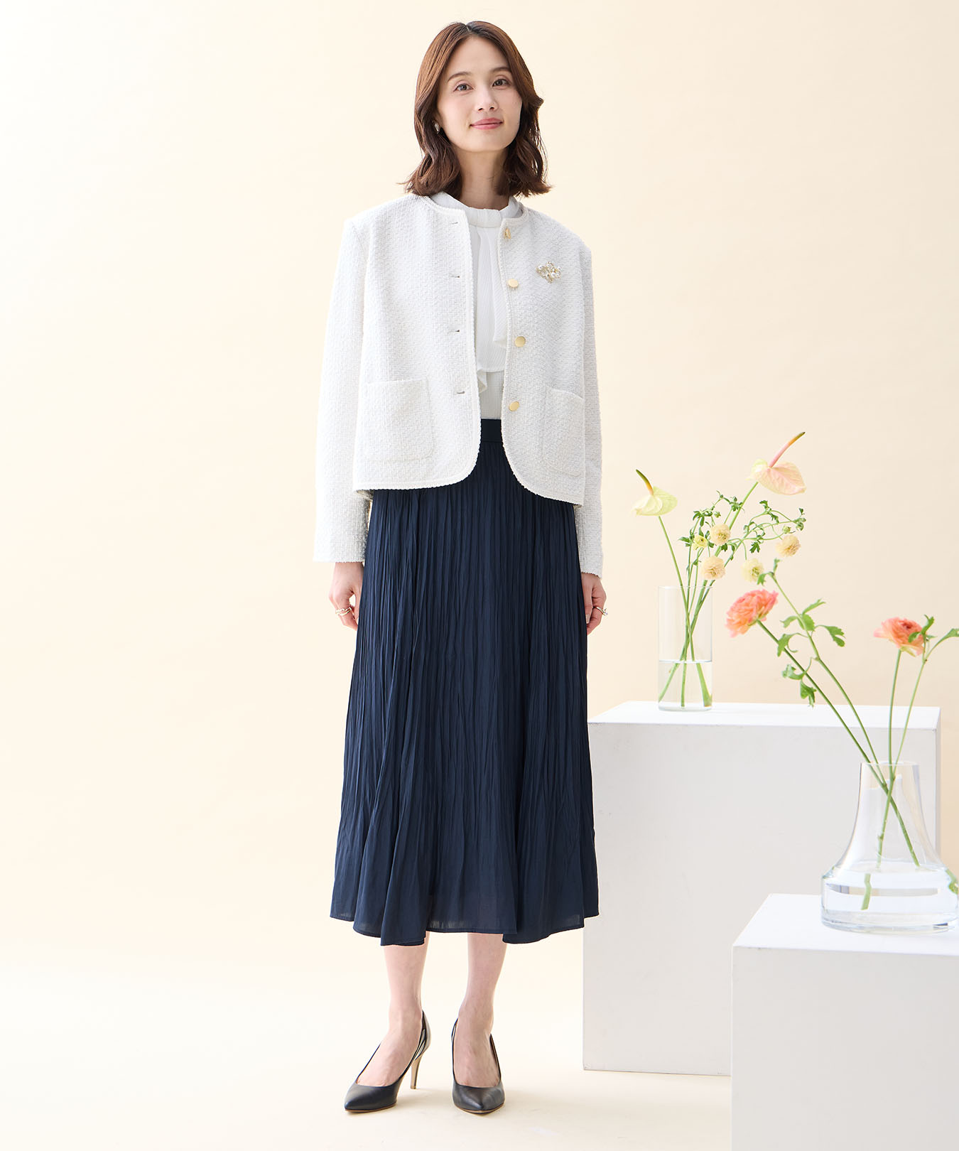 オフホワイトのツイードジャケット・ホワイトのジャガードボウタイブラウス・ネイビーのプリーツスカート・パールモチーフブローチを着用した女性の画像--1