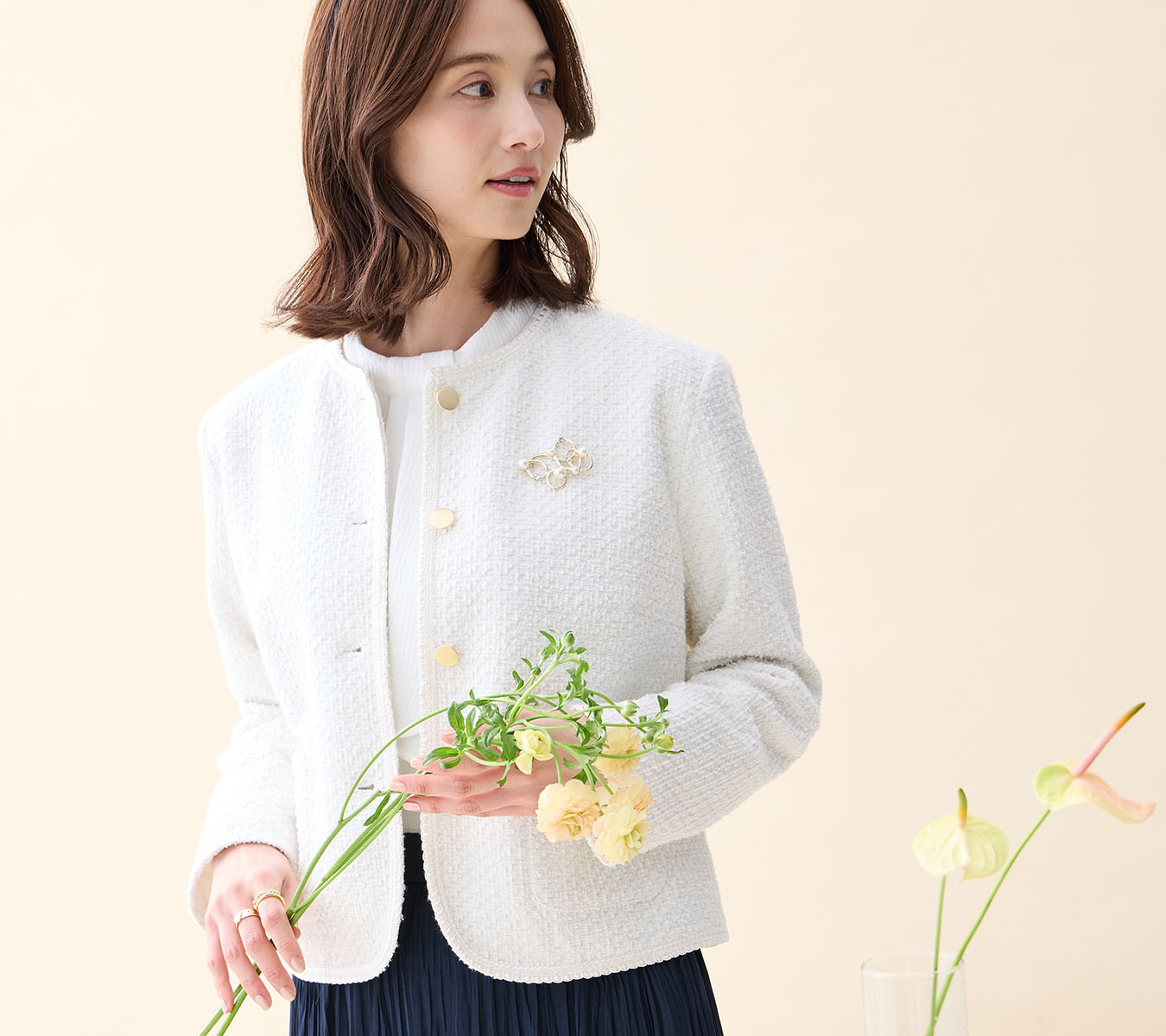 オフホワイトのツイードジャケット・ホワイトのジャガードボウタイブラウス・ネイビーのプリーツスカート・パールモチーフブローチを着用した女性の画像--2