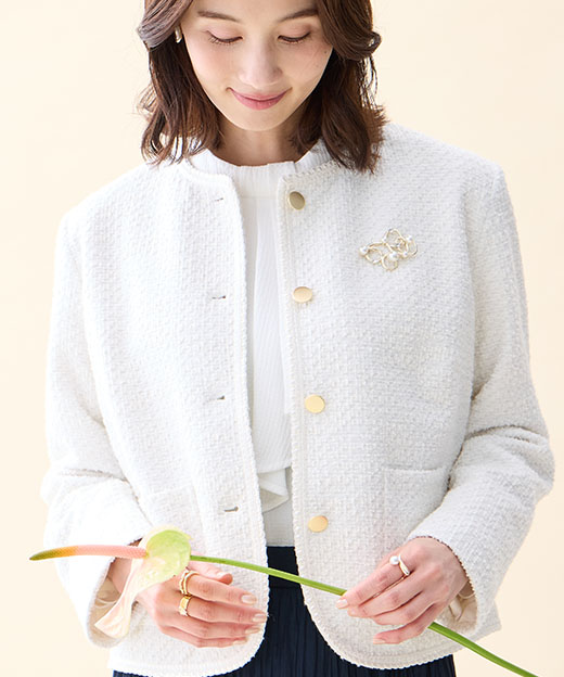 オフホワイトのツイードジャケット・ホワイトのジャガードボウタイブラウス・ネイビーのプリーツスカート・パールモチーフブローチを着用した女性の画像（ポイント）