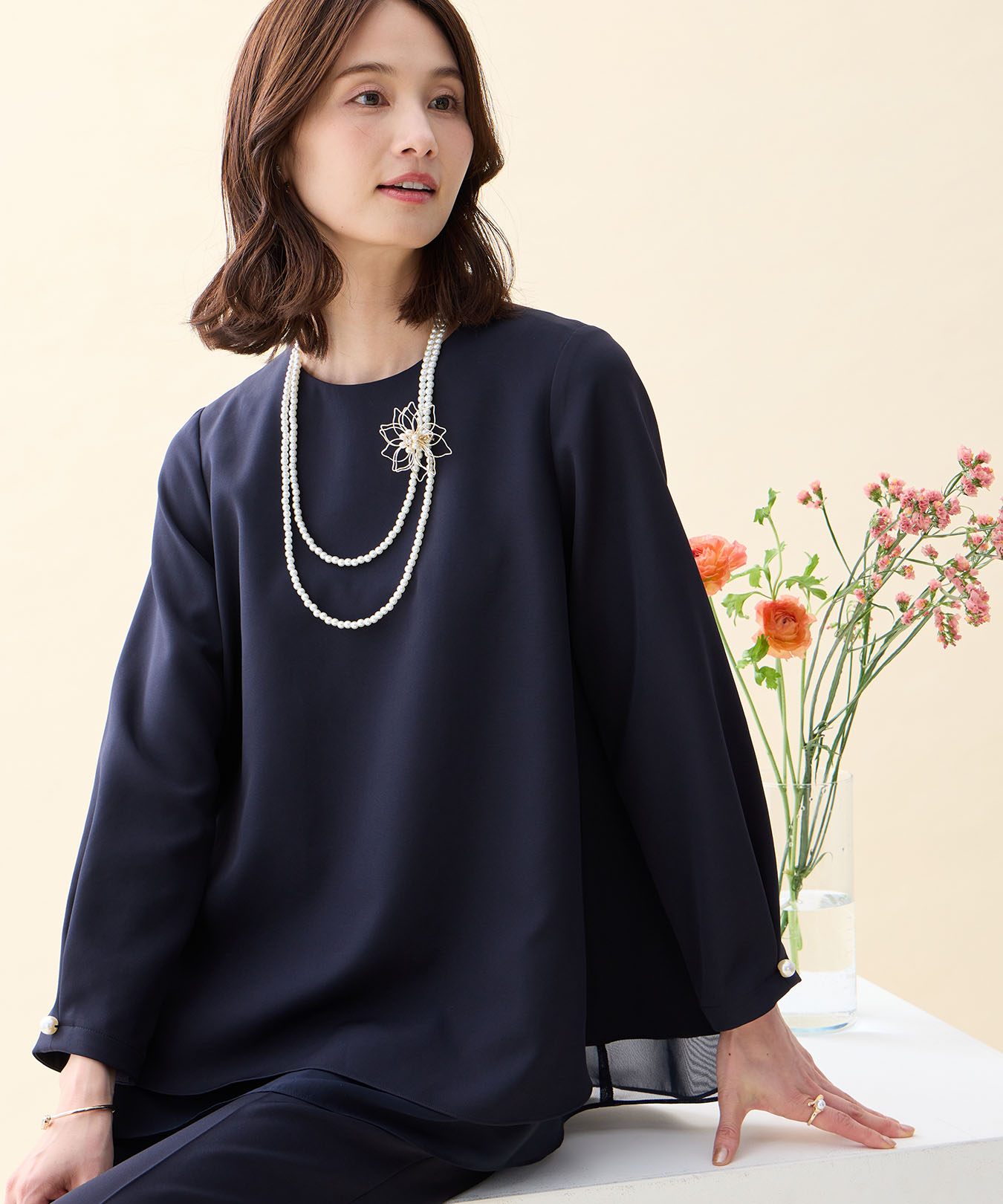 ネイビーのシフォンレイヤードブラウス・ネイビーのストレートパンツ・ブローチ付きネックレスを着用した女性の画像