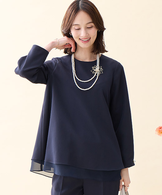 ネイビーのシフォンレイヤードブラウス・ネイビーのストレートパンツ・ブローチ付きネックレス・ベージュのトートバッグを着用した女性の画像（ポイント）