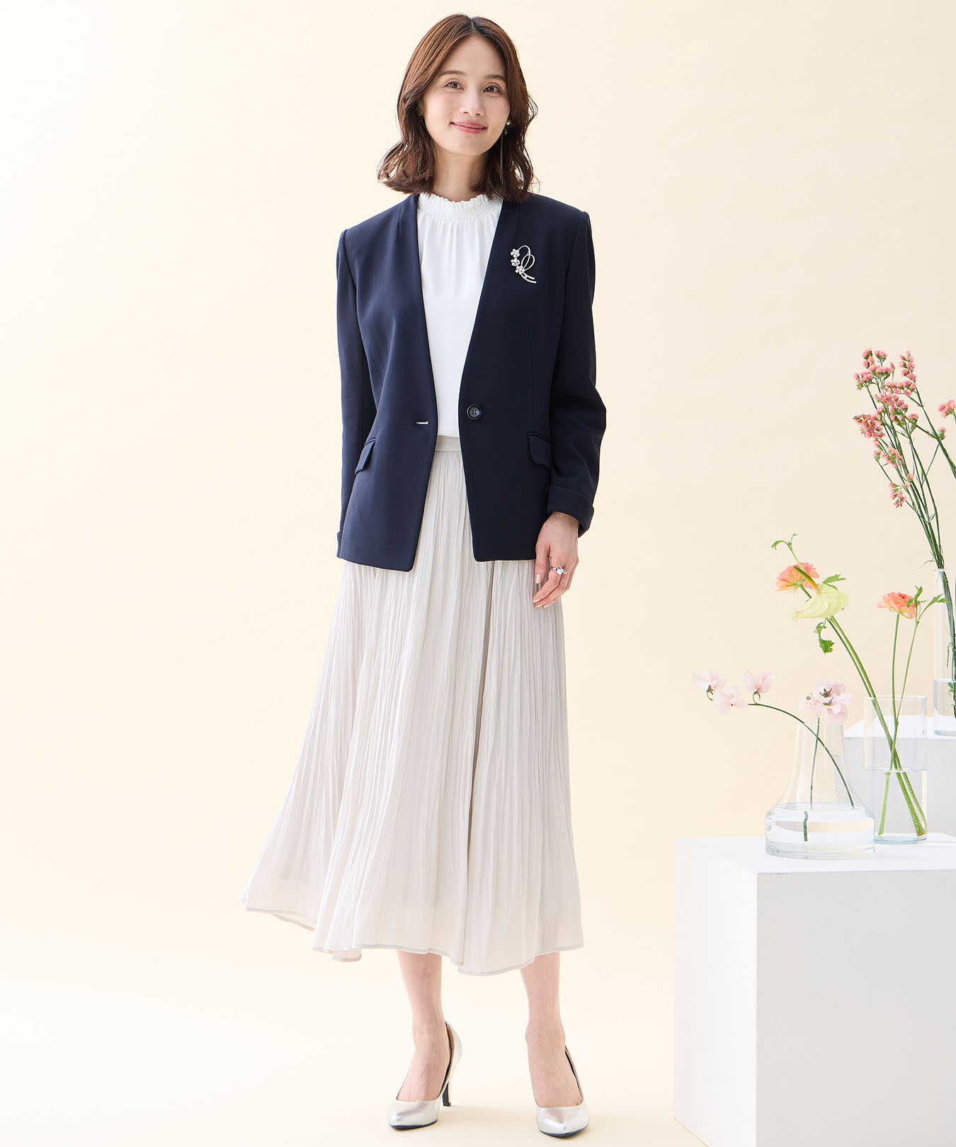 ネイビーの【アルファブリック】カラーレスジャケット・オフホワイトのフリルブラウス・ホワイトのプリーツスカート・リボンモチーフブローチを着用した女性の画像