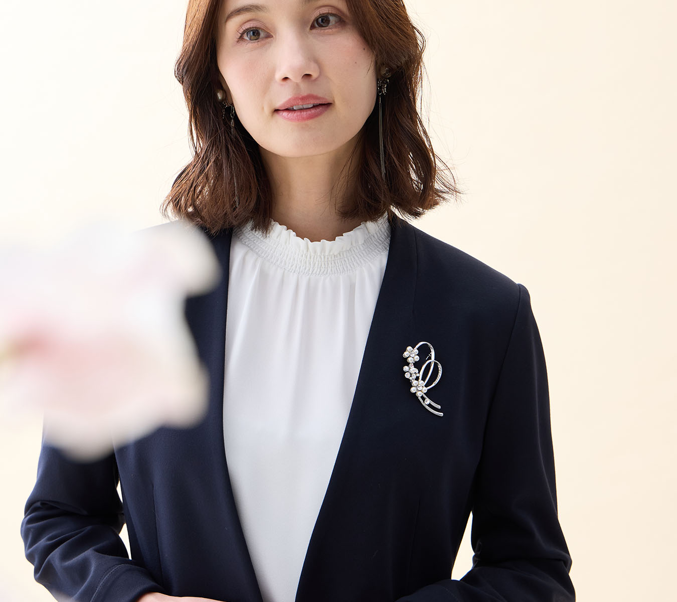 ネイビーの【アルファブリック】カラーレスジャケット・オフホワイトのフリルブラウス・リボンモチーフブローチを着用した女性の画像