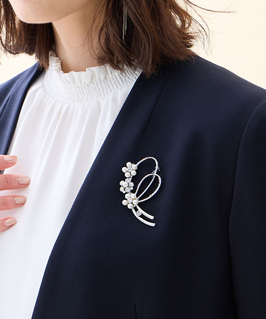 ネイビーの【アルファブリック】カラーレスジャケット・オフホワイトのフリルブラウス・リボンモチーフブローチを着用した女性の画像（ポイント）--1