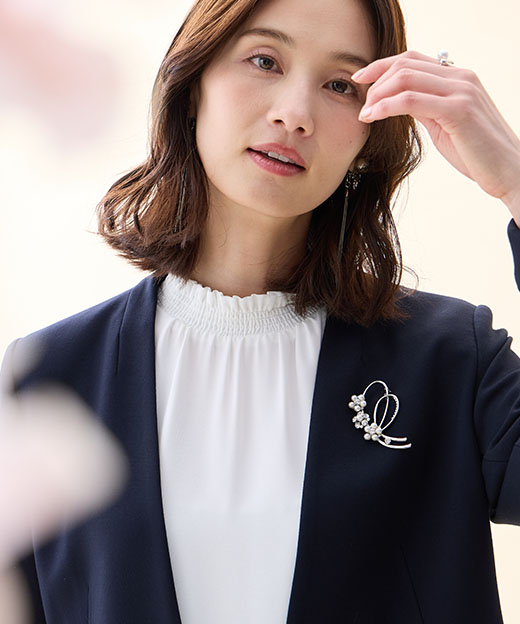 ネイビーの【アルファブリック】カラーレスジャケット・オフホワイトのフリルブラウス・リボンモチーフブローチを着用した女性の画像（ポイント）--2