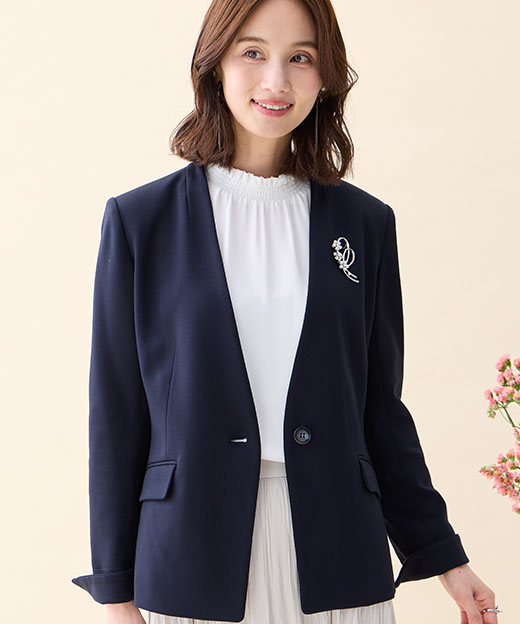 ネイビーの【アルファブリック】カラーレスジャケット・オフホワイトのフリルブラウス・ホワイトのプリーツスカート・リボンモチーフブローチを着用した女性の画像（ポイント）