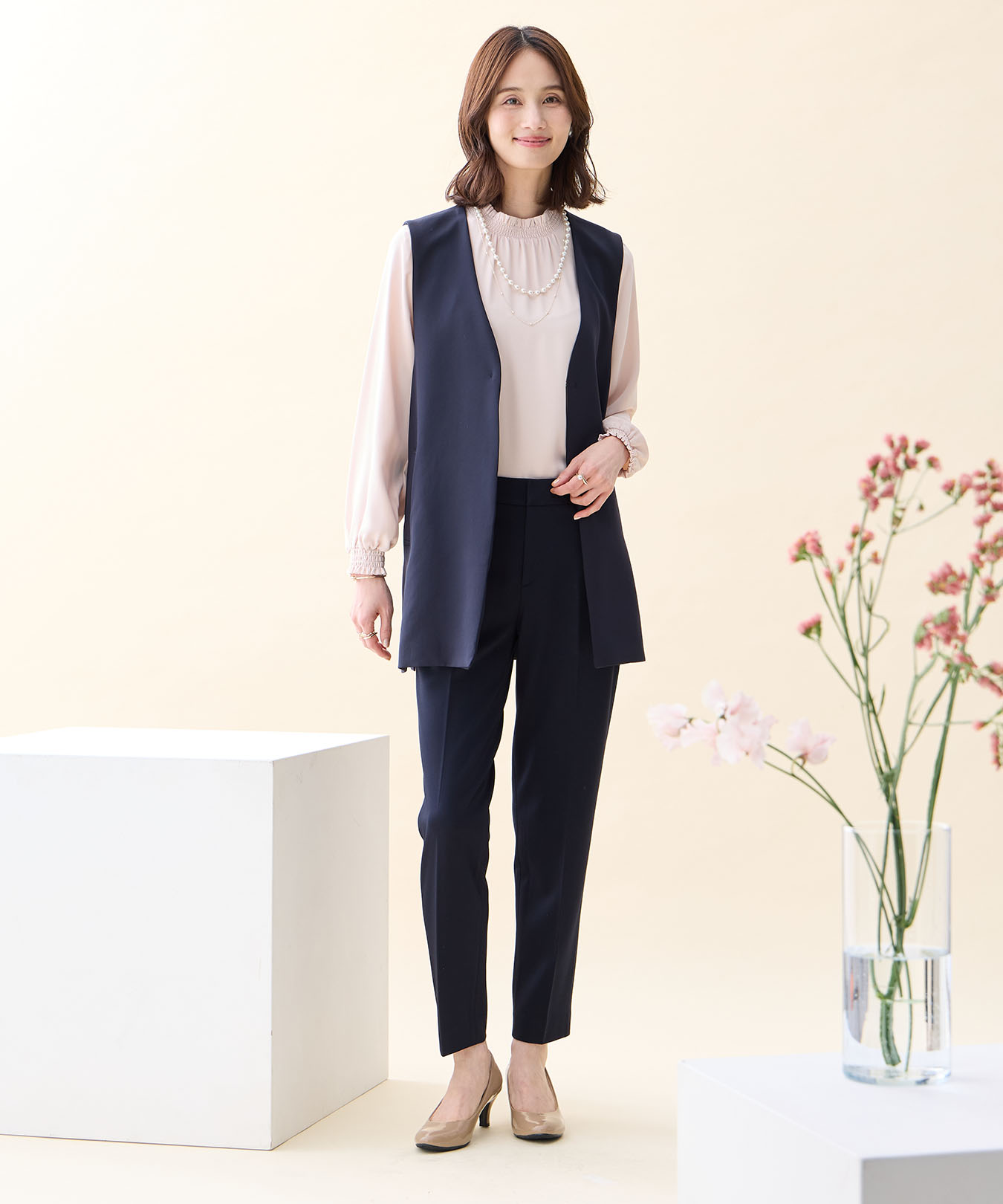 ネイビーのジレジャケット・ピンクのフリルブラウス・ネイビーの【アルファブリック】テーパードパンツ・パールネックレスを着用した女性の画像--1