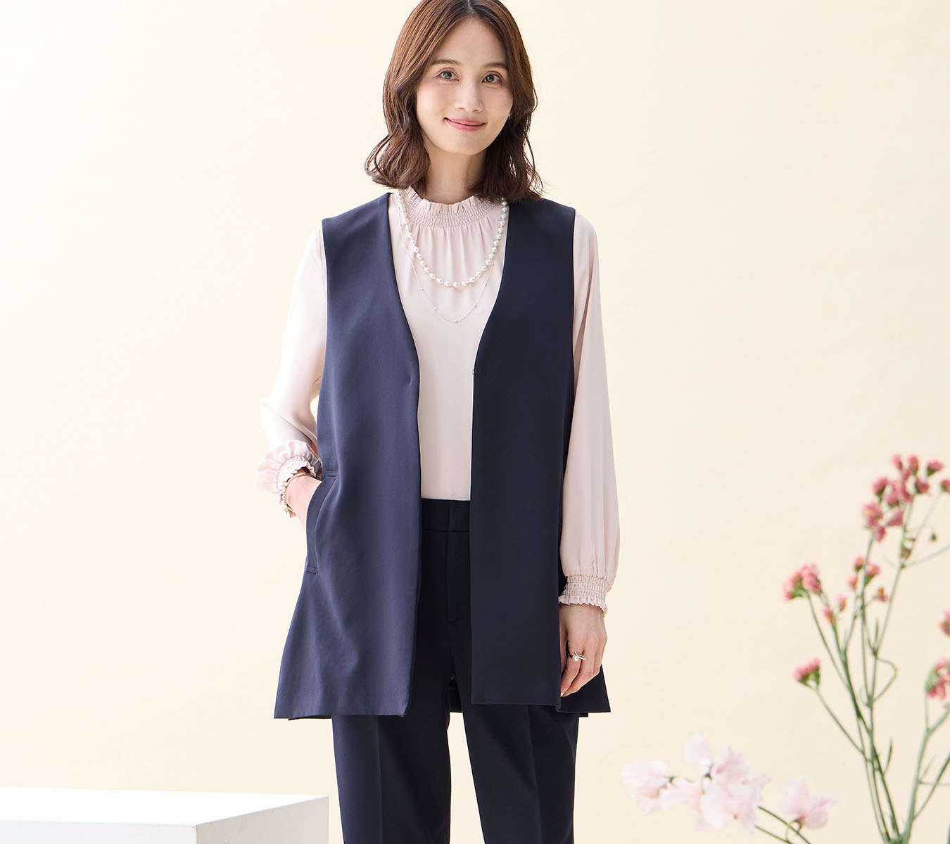 ネイビーのジレジャケット・ピンクのフリルブラウス・ネイビーの【アルファブリック】テーパードパンツ・パールネックレスを着用した女性の画像--2