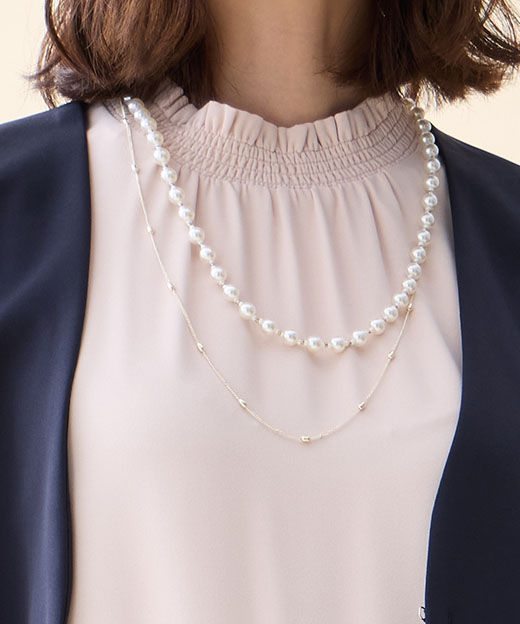 ネイビーのジレジャケット・ピンクのフリルブラウス・ネイビーの【アルファブリック】テーパードパンツ・パールネックレスを着用した女性のアップの画像（ポイント）