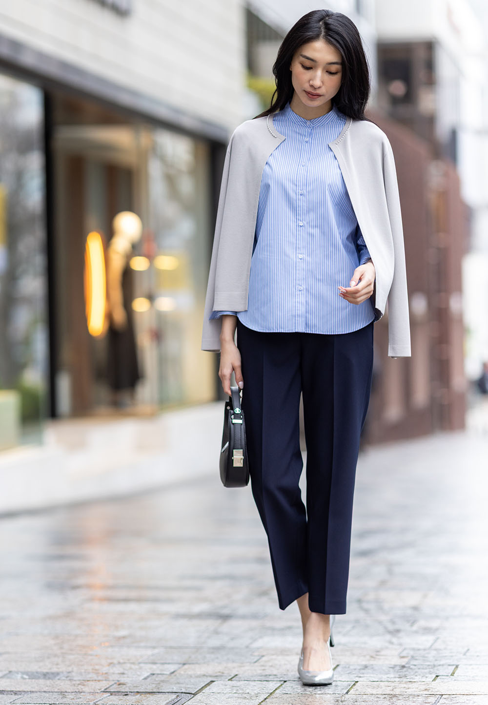 ライトグレーのパールボタンニットカーディガン・ブルーのバンドカラーストライプシャツ・ネイビーのストレッチ テーパードパンツを着用した女性の画像