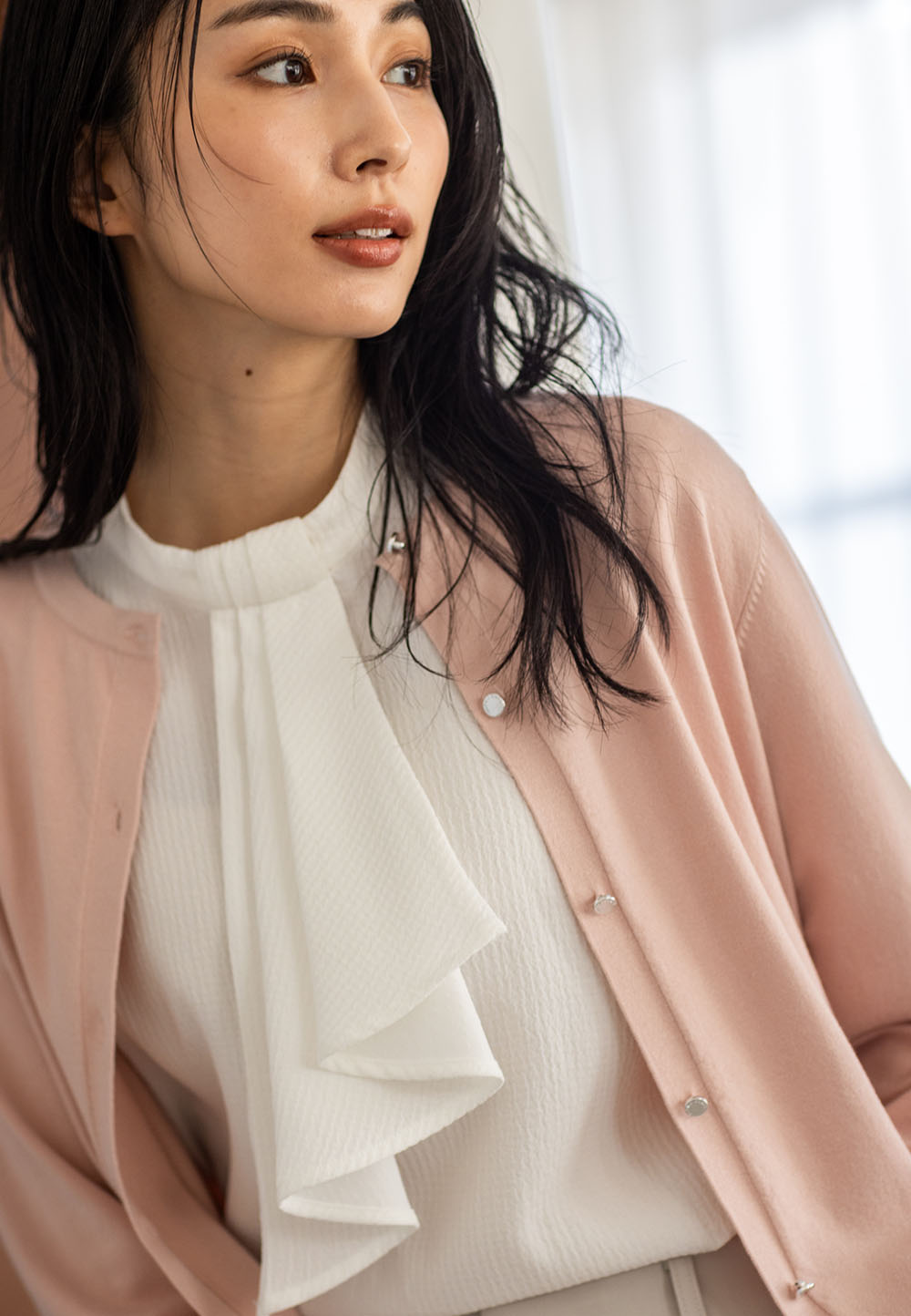 ピンクのクルーネックニットカーディガン・オフホワイトのジャガードボウタイブラウス・アイボリーのストレッチ テーパードパンツを着用した女性のアップの画像