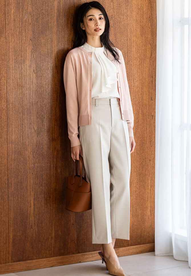 ピンクのクルーネックニットカーディガン・オフホワイトのジャガードボウタイブラウス・アイボリーのストレッチ テーパードパンツを着用した女性の画像