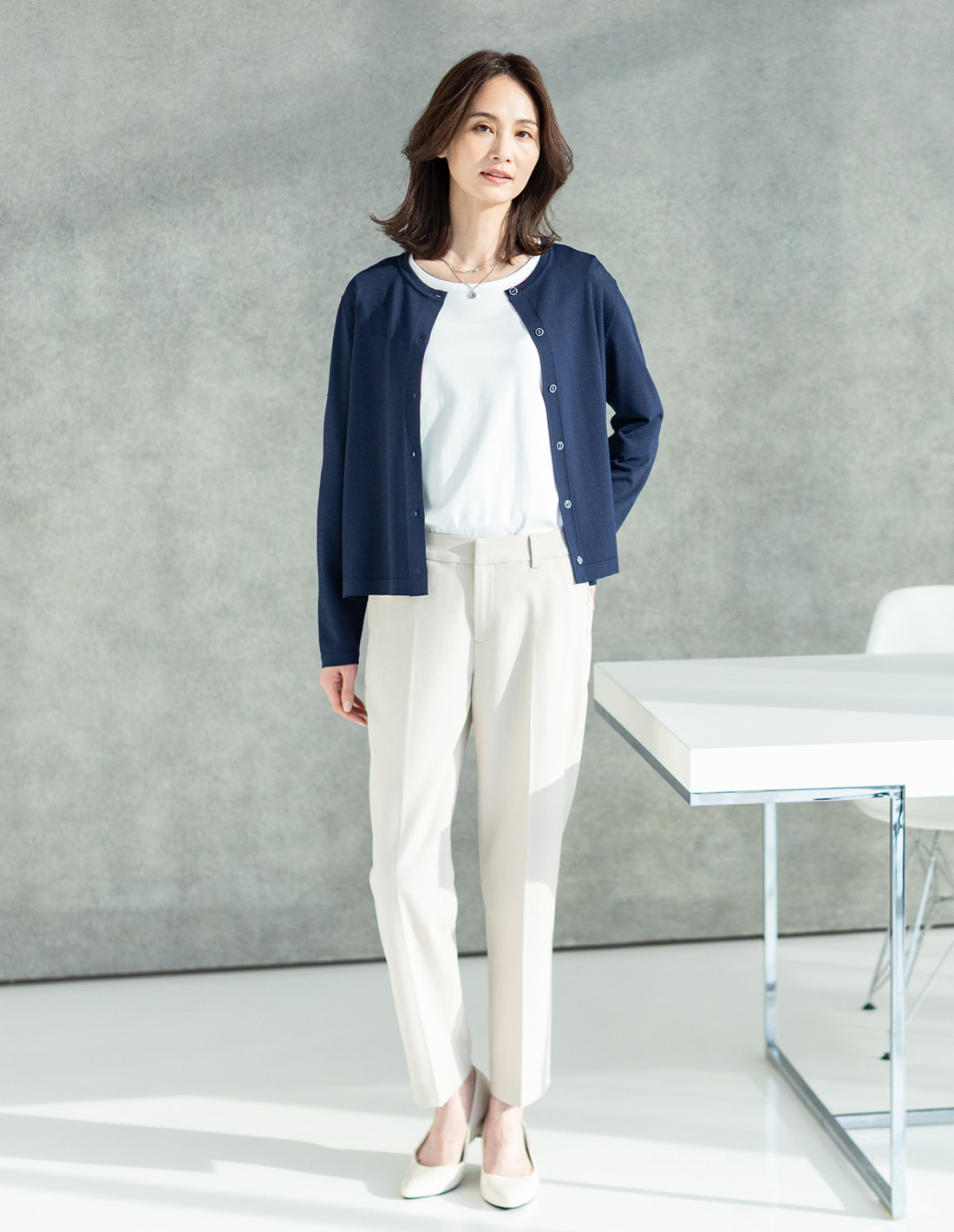 ライトベージュのストレッチテーパードパンツ・ネイビーのクルーネックカーディガン・オフホワイトのレースドッキングプルオーバー・２連モチーフトップネックレスを着用した女性の画像