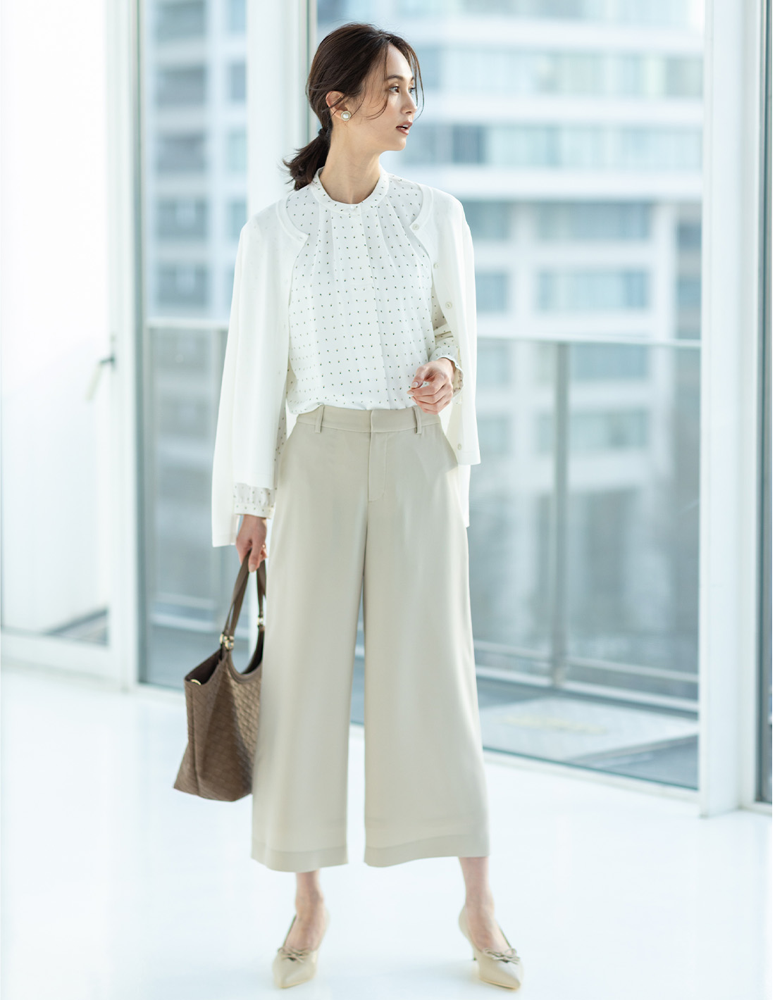 オフホワイトのクルーネックカーディガン・オフホワイトの幾何プリントジョーゼットブラウス・ベージュのストレッチジョーゼットワイドパンツ【セットアップ対応】・編み込み風合皮トートバッグを着用した女性の画像