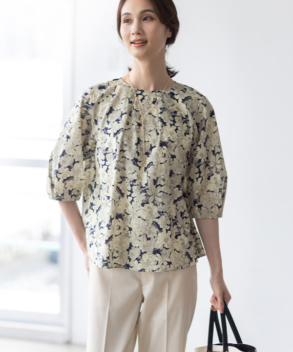 リバティプリントタックスリーブブラウス、バーズアイストレッチクロップドパンツ、ボールチェーンネックレスを着用した女性の画像（アンカーリンク）