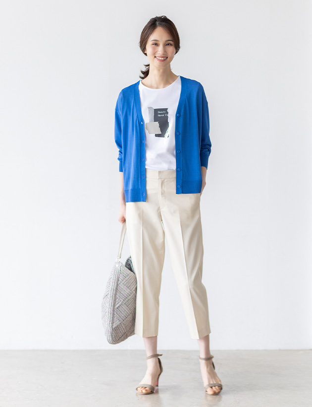 ベージュのバーズアイストレッチクロップドパンツ・ブルーのVネックカーディガン・ホワイトのグラフィックロゴTシャツ・リバティプリントワンショルダーバッグを着用した女性