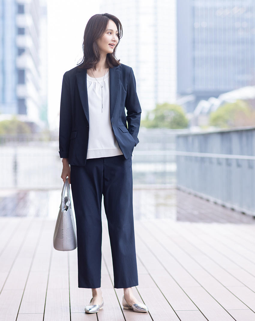 ネイビーのストレッチストレートパンツ・ネイビーのストレッチテーラードジャケット・ホワイトのフロント切替ニットプルオーバー・グレーのパンチング合皮2WAYトートバッグを着用した女性