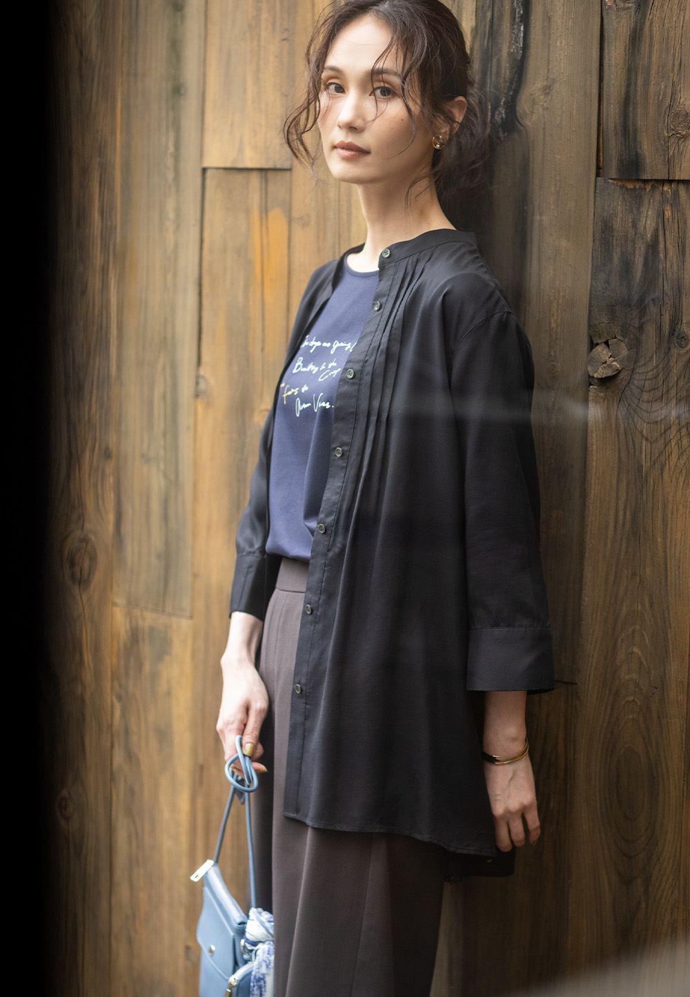 タックブラウスの羽織り・ブラウンのストレートパンツを着用し、左斜め前を向いて壁の前に立つ女性