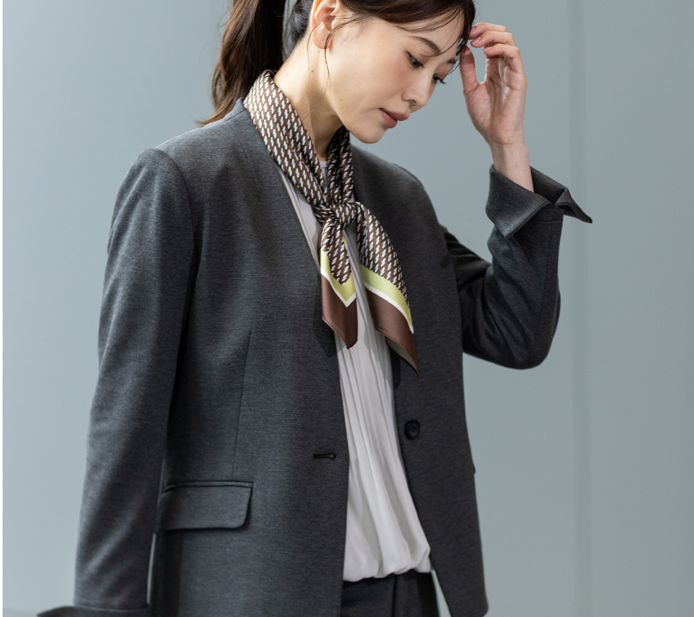 グレーのジャケット・シフォンブラウス・スカーフを着用し、下を向いている女性