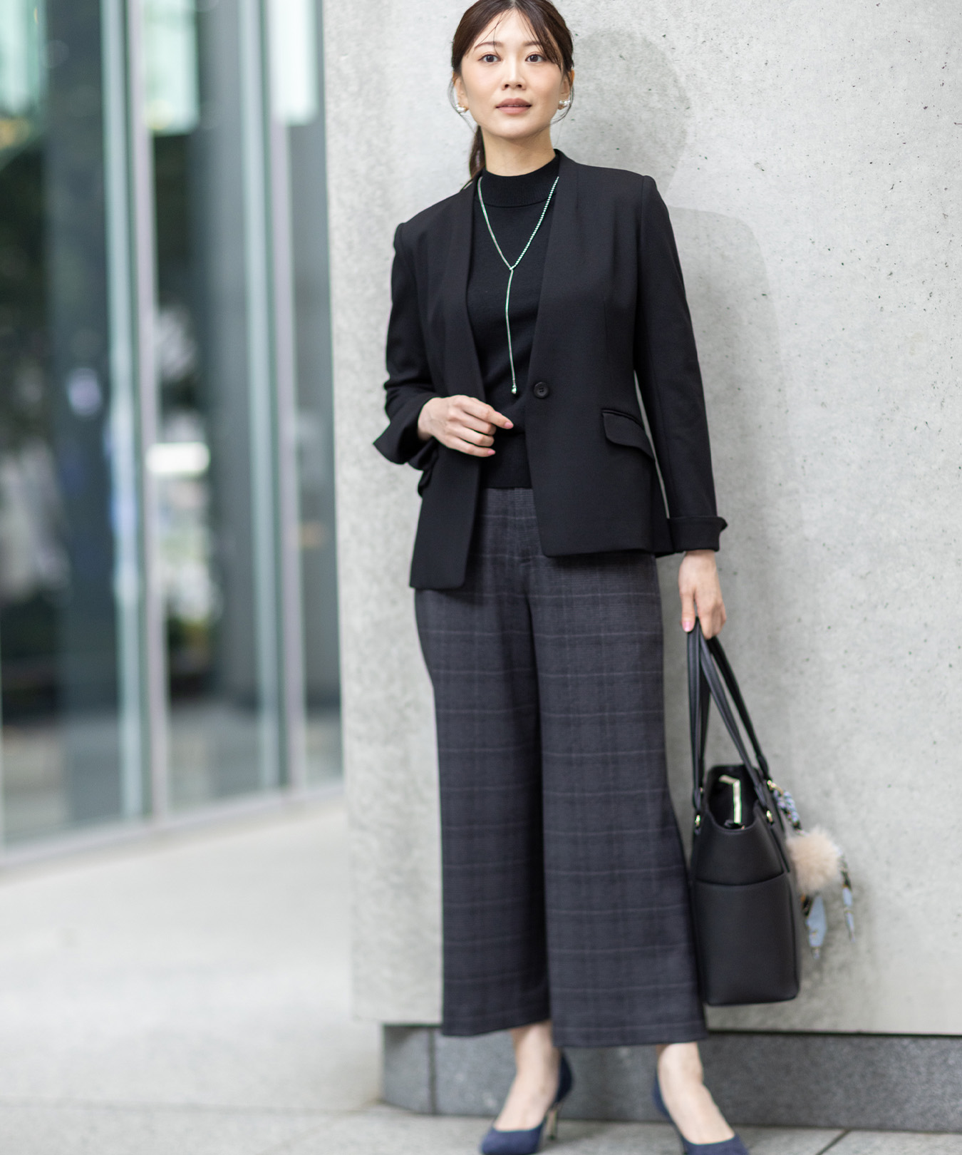 グレーのワイドパンツ・ブラックのジャケットを着用し、壁の前に立っている女性