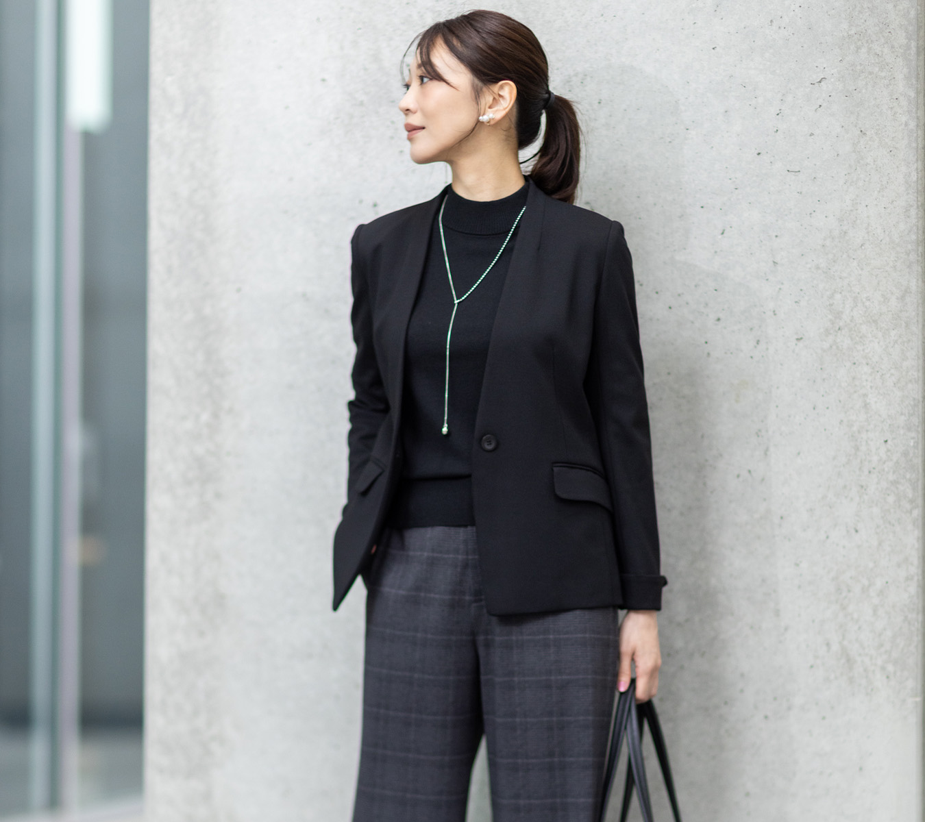 グレーのワイドパンツ・ブラックのトップス・ブラックのジャケットを着用し、壁の前に立っている女性