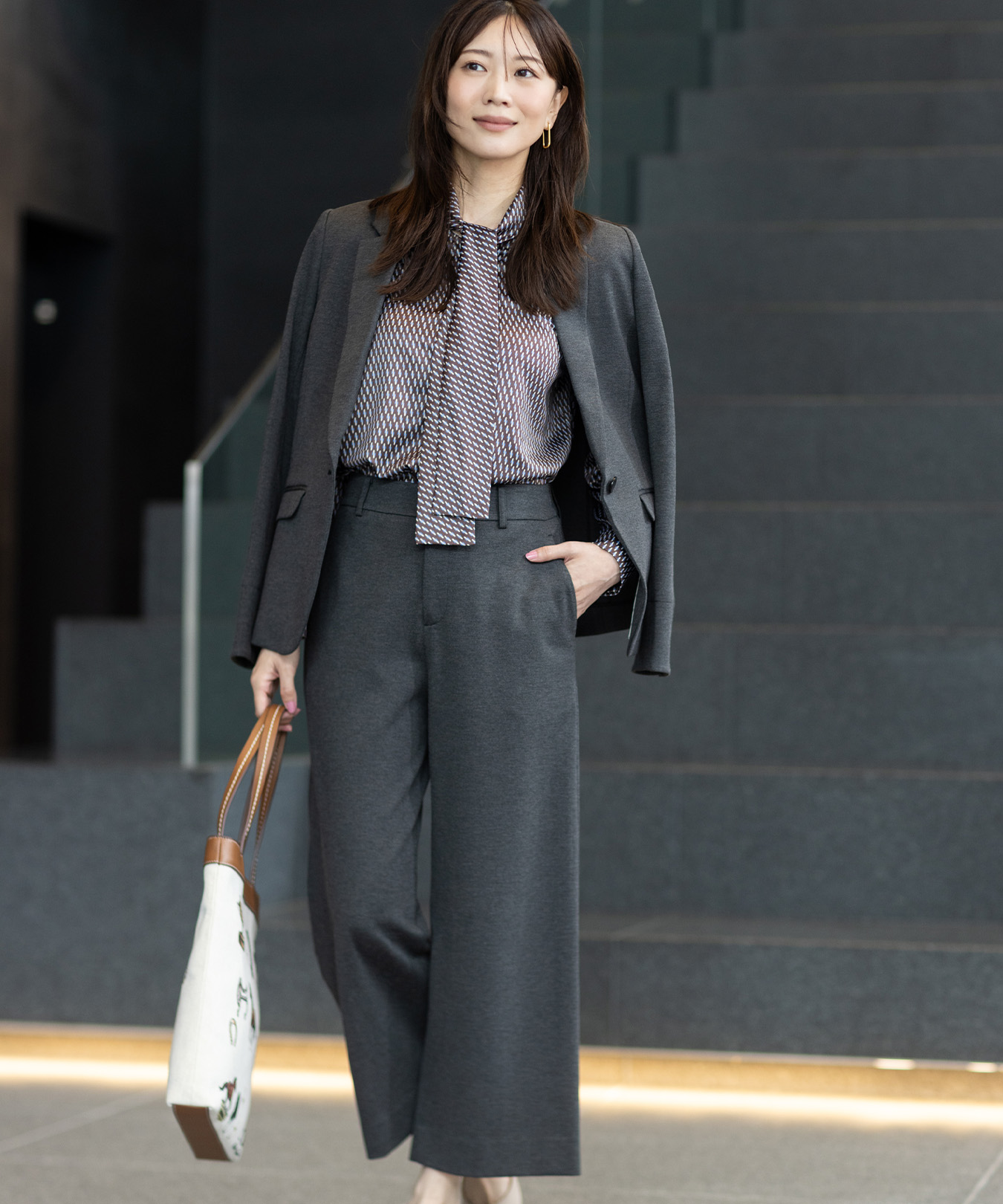 グレーのジャケット・グレーのワイドパンツ・ボウタイブラウスを着用し、立っている女性