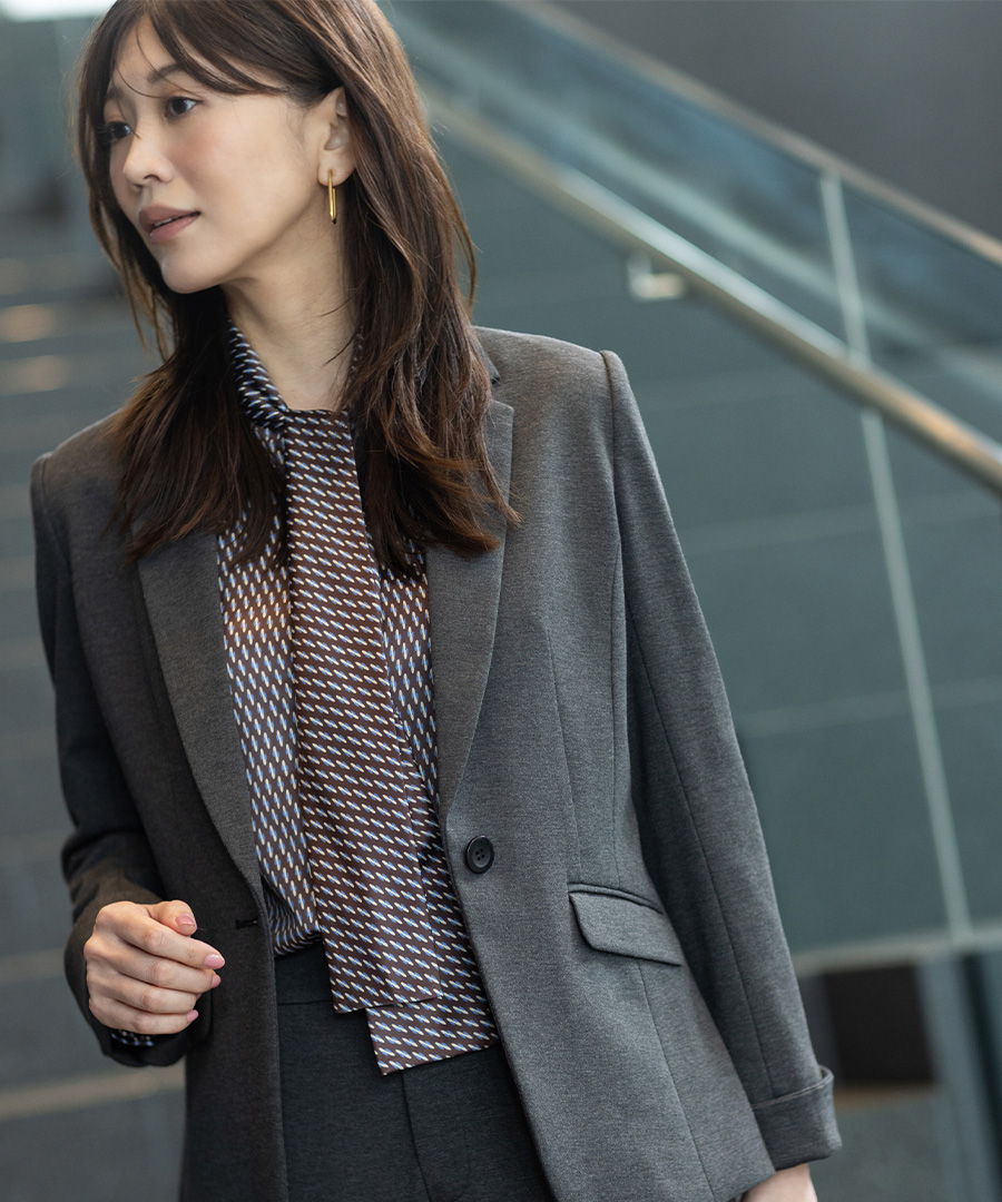 グレーのジャケット・グレーのワイドパンツ・ボウタイブラウスを着用し、左を向いた女性のアップ