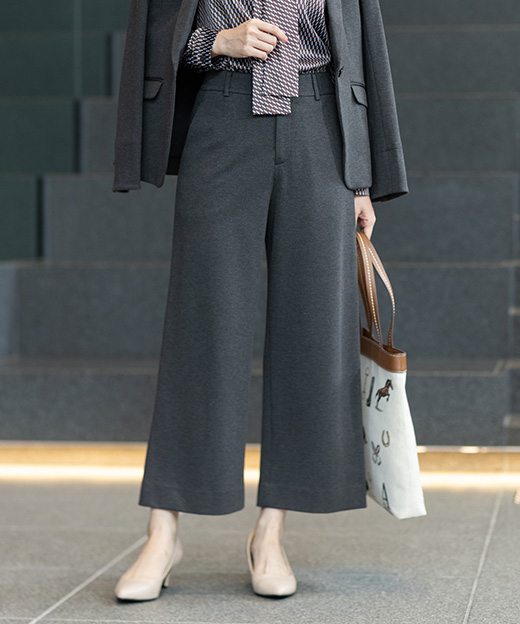 グレーのジャケット・グレーのワイドパンツ・ボウタイブラウスを着用し、立っている女性の足元