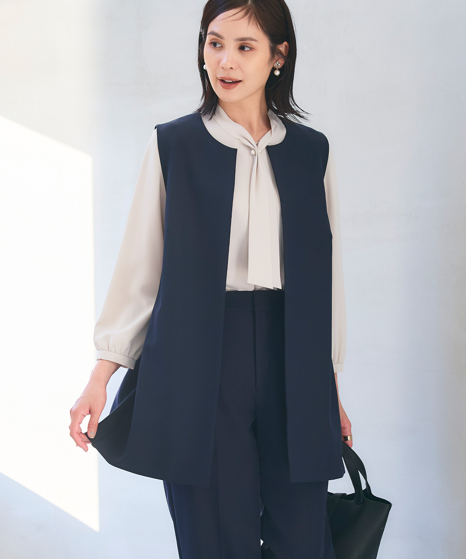 ネイビーの《洗える・シワになりにくい》正解ロングジレ・ネイビーの《洗える・シワになりにくい》正解ワイドパンツ・オフホワイトの《シワになりにくい》シルキーサテンパールボウタイブラウスを着用した女性の画像（スライド）