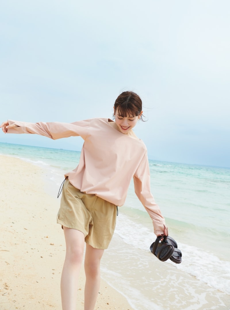 ドロストヘムラッシュガードとラッシュガードショートパンツを着用した女性がサンダルを片手に持って砂浜を歩いている写真