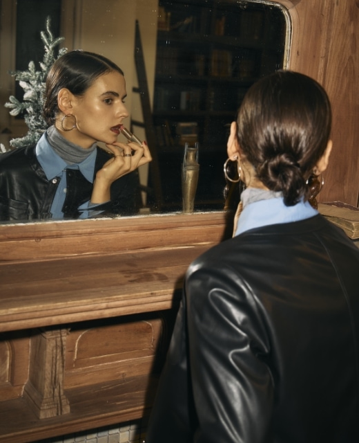 黒のフェイクレザーショートジャケット・ブルーのコンフォータブルシャツ・グレーのメリノウールシアータートルを着用した女性が鏡の前で化粧直しをするビジュアル