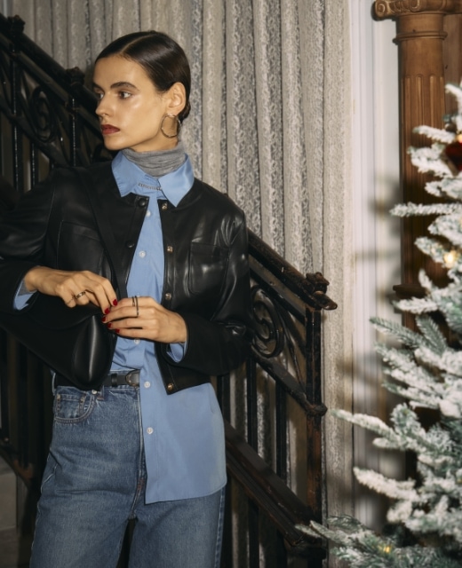 黒のフェイクレザーショートジャケット・ブルーのコンフォータブルシャツ・グレーのメリノウールシアータートル・ミッドブルーの【NEEDBY】ANNIEのフレアデニムを着用した女性のビジュアル。シャツの裾を右側だけパンツインした着こなし。