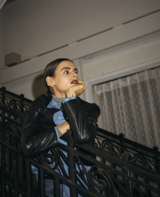 黒のフェイクレザーショートジャケット・ブルーのコンフォータブルシャツ・グレーのメリノウールシアータートルを着用した女性が階段の手すりで頬杖をついているビジュアル