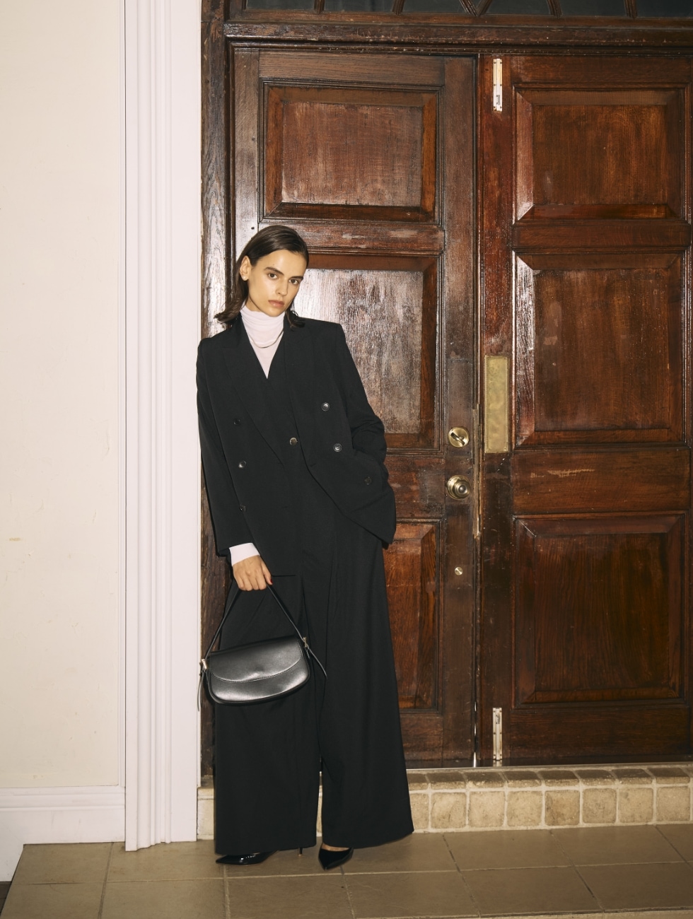 黒のピークドラペルダブルジャケット【CLEAN MOTION】とウォッシャブルオールインワン【CLEAN MOTION】・オフ白のメリノウールシアータートルを着用した女性が黒の【AULENTTI】レザーショルダーBAGを持ってドアの前でポーズをとるビジュアル
