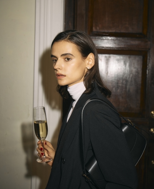 黒のピークドラペルダブルジャケット【CLEAN MOTION】・オフ白のメリノウールシアータートルを着用した女性が片手にシャンパンを持つバストアップのビジュアル