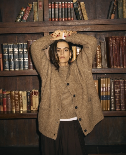 ブラウンのアルパカシャギーニットカーディガンとアルパカシャギーニットプルオーバー、黒のウールライクボリュームドロストパンツを着用した女性が洋書の本棚の前でポーズをとるビジュアル