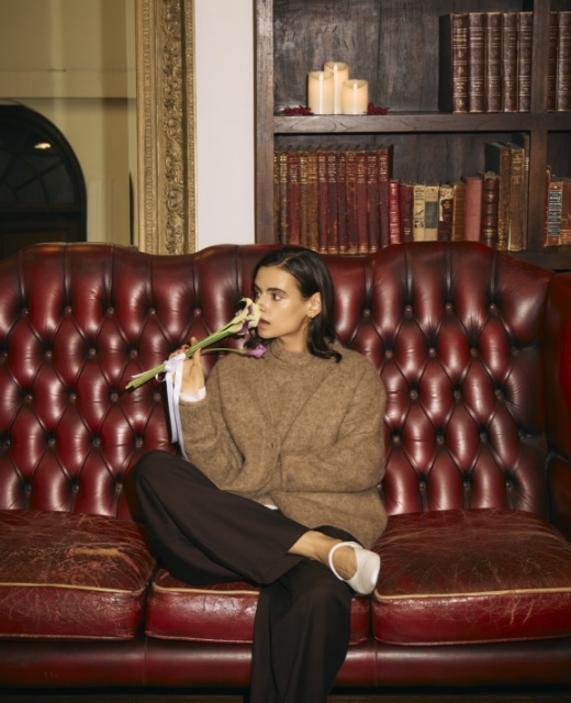 ブラウンのアルパカシャギーニットカーディガンとアルパカシャギーニットプルオーバー、黒のウールライクボリュームドロストパンツを着用した女性がソファに座り、花束の香りを嗅ぐビジュアル