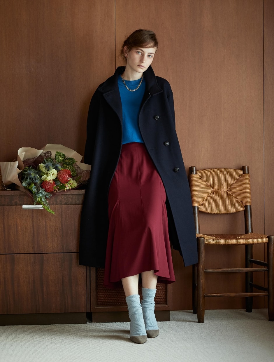 コートとニットとスカートを着用した女性が花束の横に立っている写真