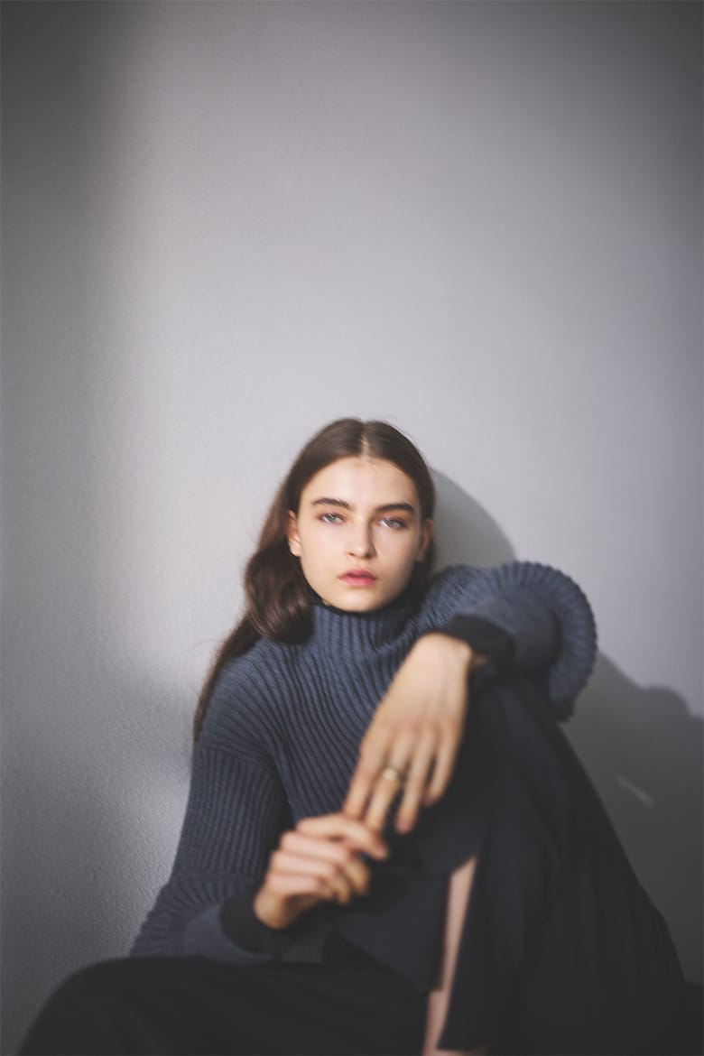 紺色のプルオーバー、黒のパンツを着用し、壁に背を当て座っている女性モデルの写真